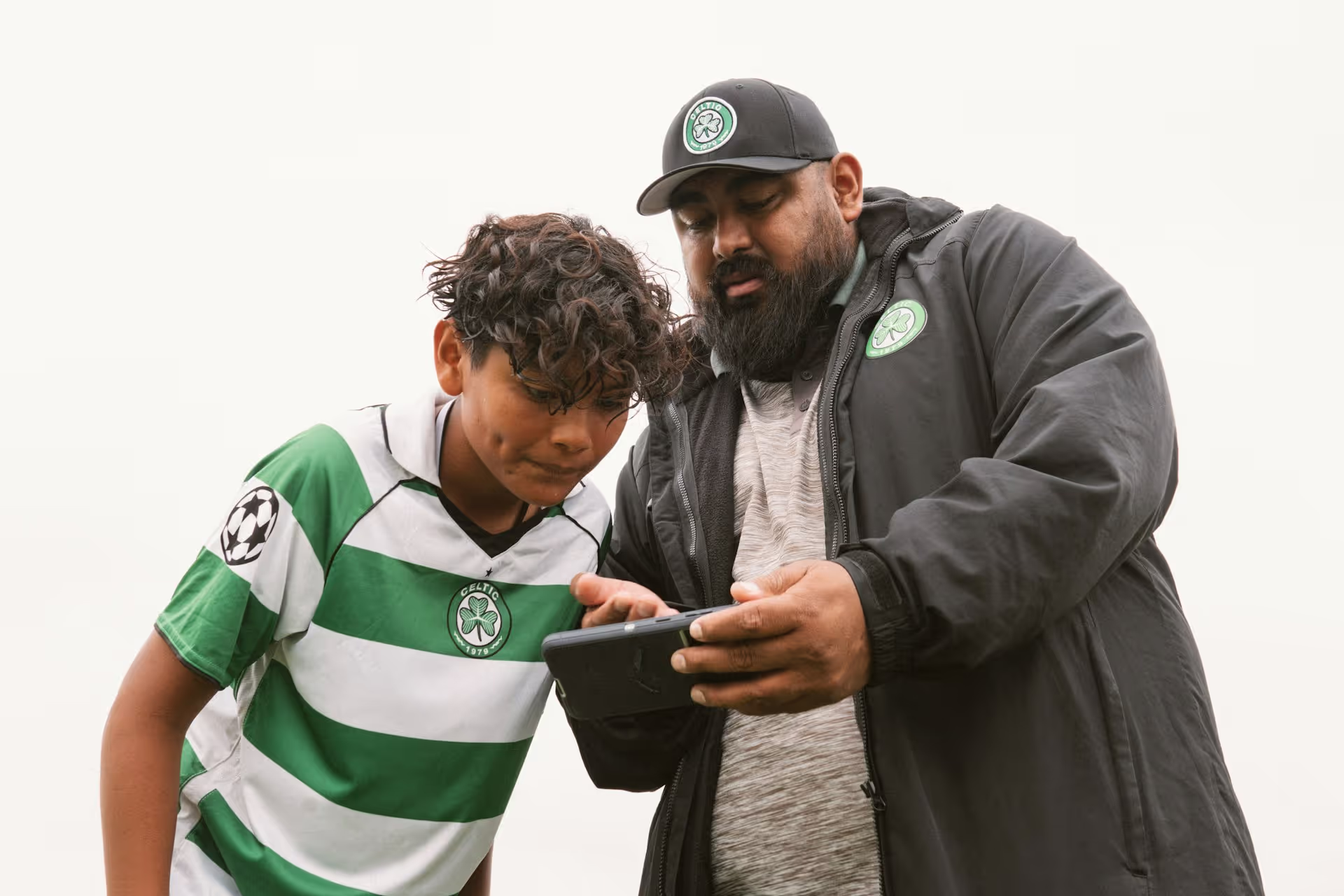 A coach showing a young soccer player something on a smartphone, both wearing Celtic sportswear.