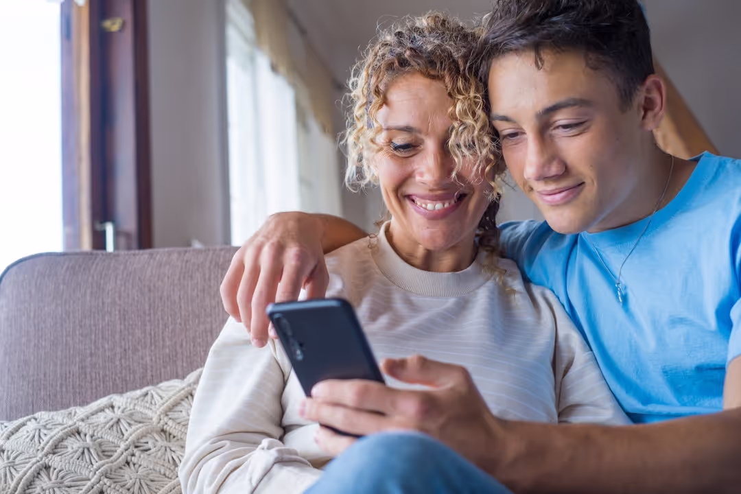 Smiling woman and young man sitting closely on a couch, looking at a smartphone together.