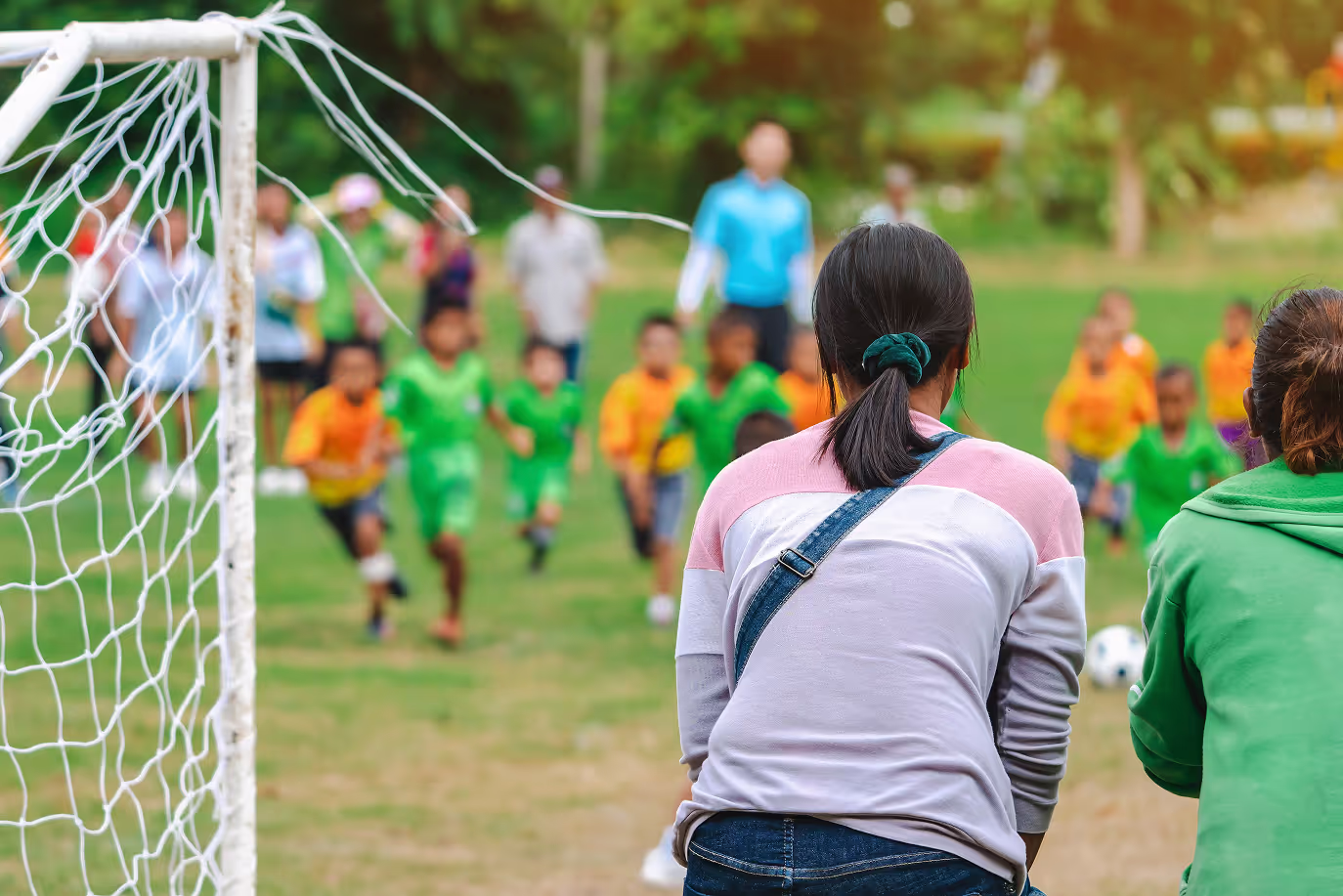 Children in green and orange uniforms running on a soccer field, viewed from behind two adults near a goalpost.