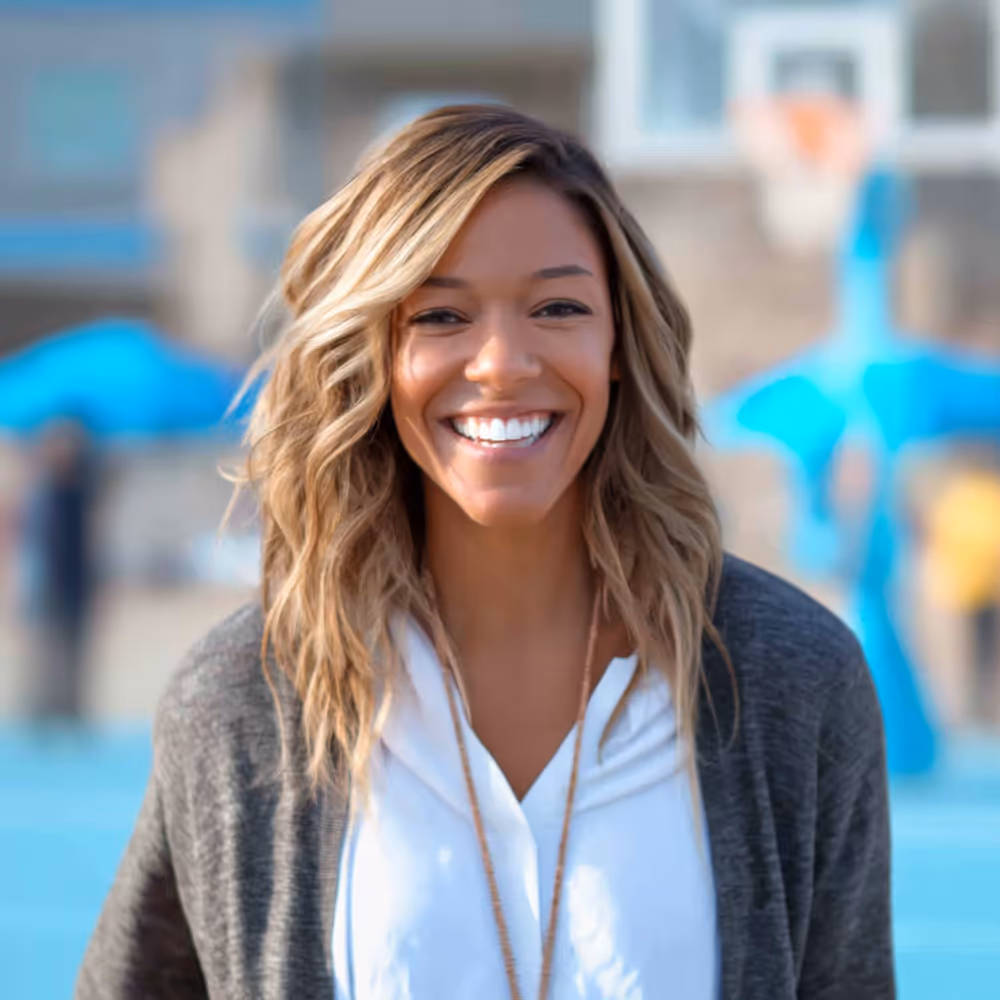 Smiling woman with wavy blonde hair wearing a white top and gray cardigan standing outdoors near a basketball court.