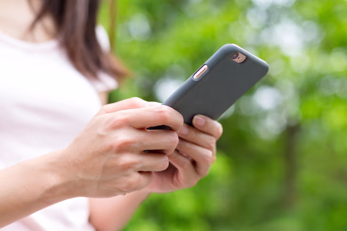 Close-up of person holding and using a smartphone with a black case outdoors with green blurred background.