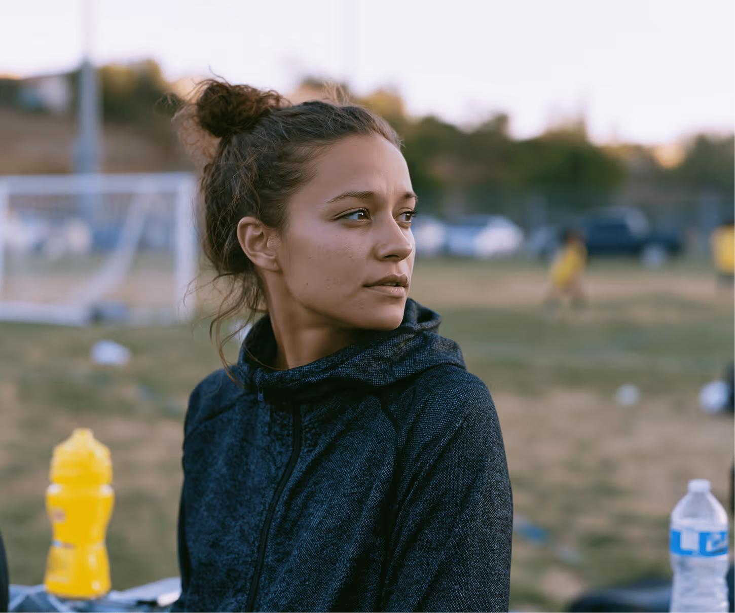 Young woman in a dark jacket with hair in a bun sitting outdoors near a soccer field.
