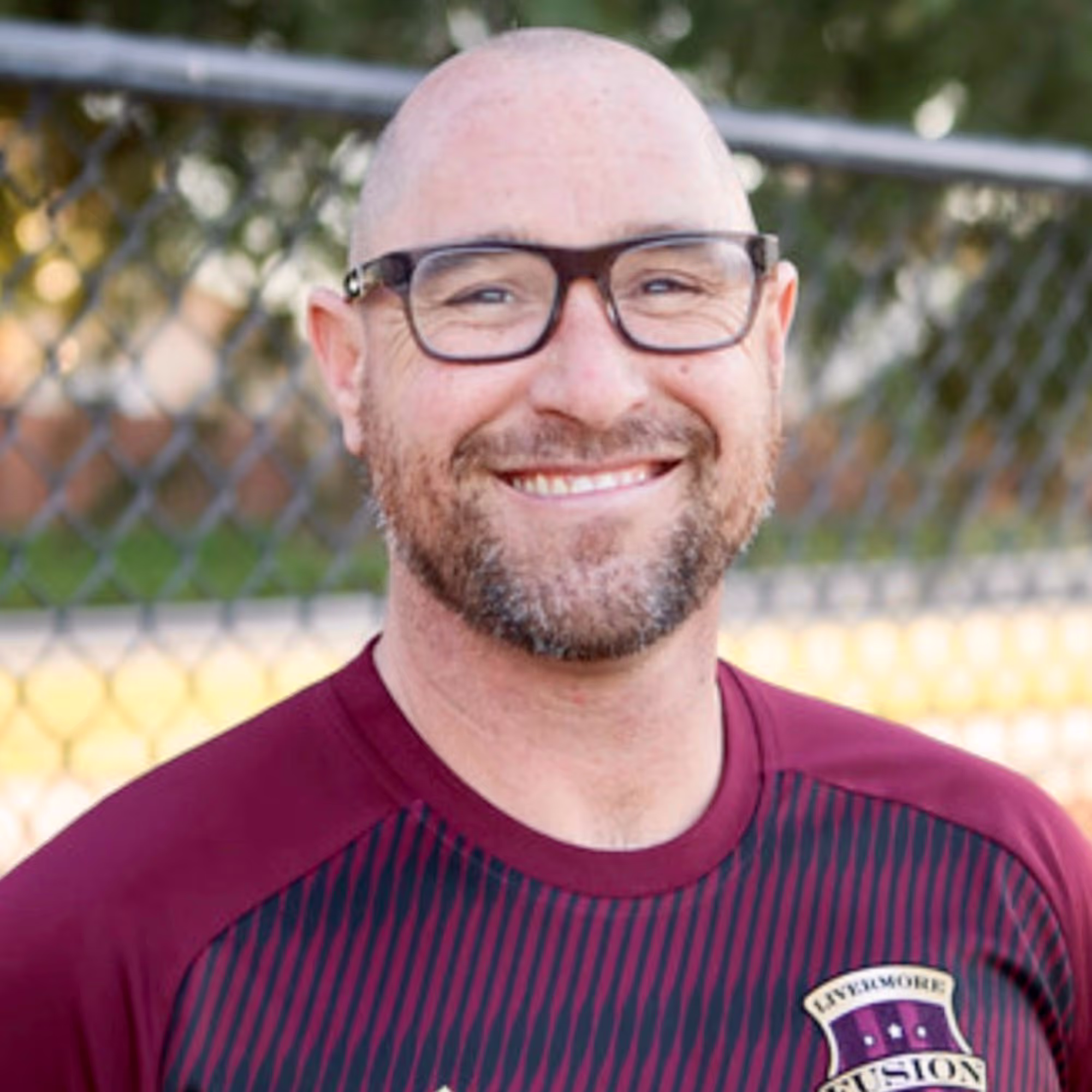 Smiling bald man with glasses and a beard wearing a maroon Livermore Fusion sports jersey.