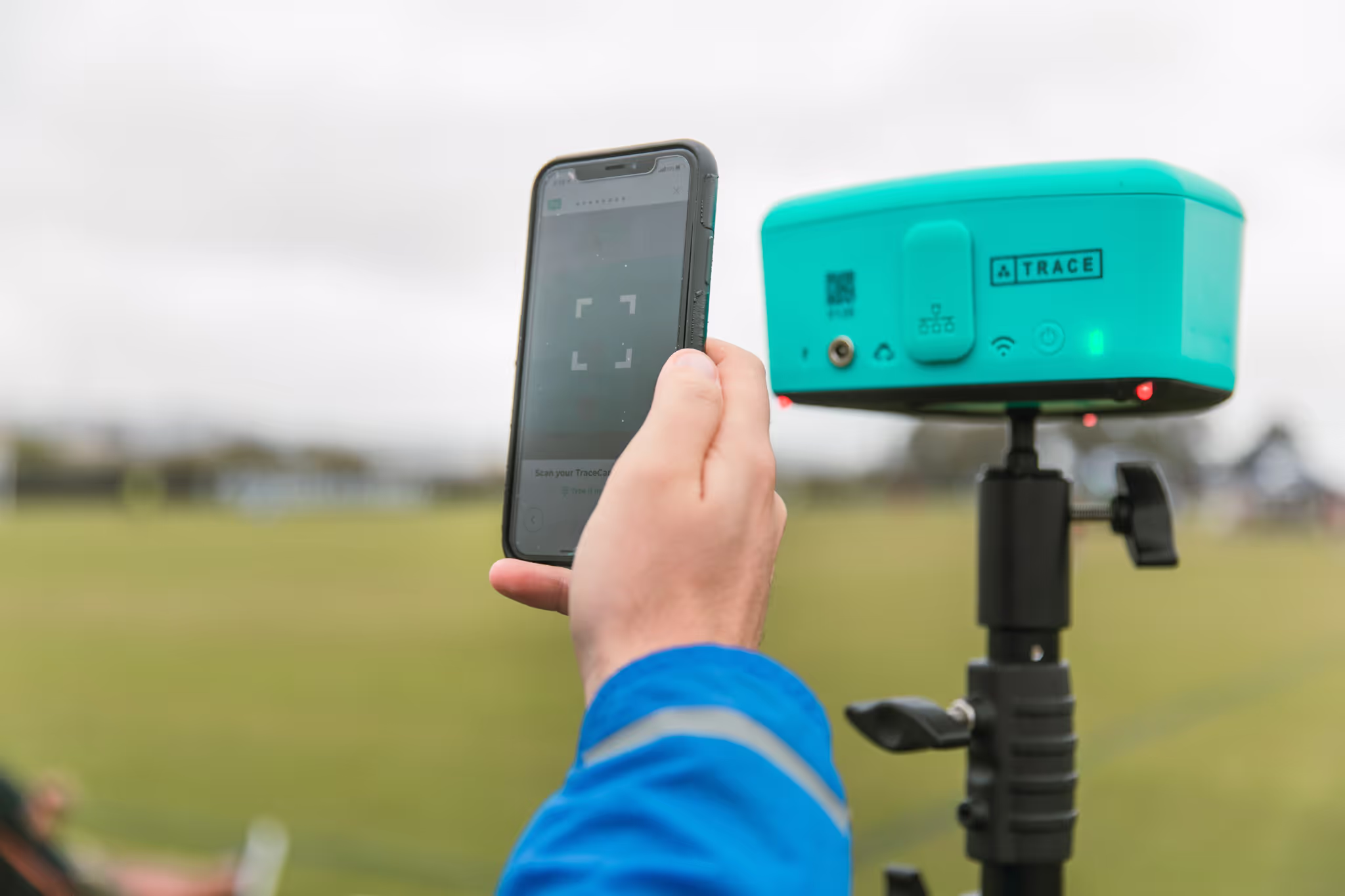 Hand holding a smartphone scanning a turquoise Trace device mounted on a tripod outdoors.