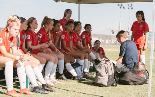 Coach Talking To Youth Team Youth girls soccer team in red kits listen to a coach kneeling in front of the sideline bench