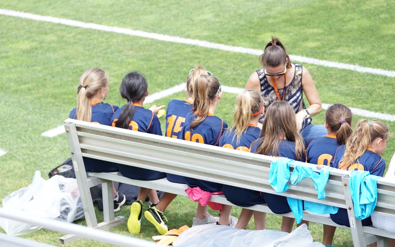 Youth Team On Sideline Bench Girls youth soccer team in navy and orange jerseys sit on a sideline bench with their coach