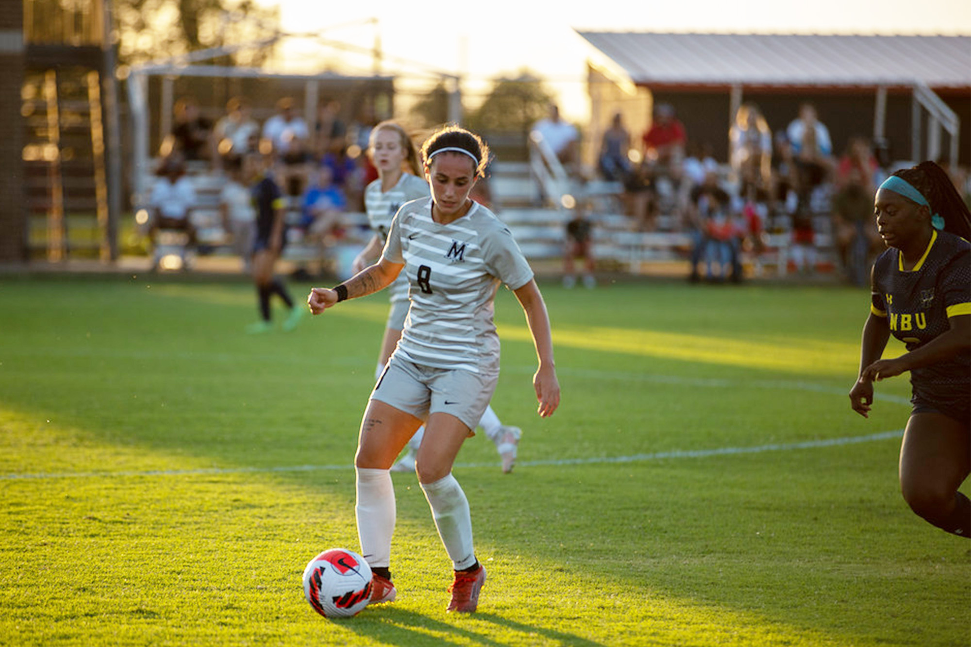 Female soccer player in a white number 8 jersey dribbles the ball at golden hour as a defender chases
