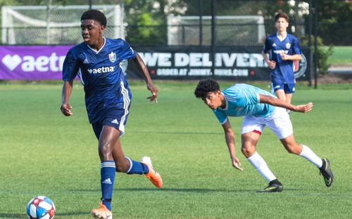 Boy in a navy Aetna-sponsored kit dribbles the ball past an opponent in teal