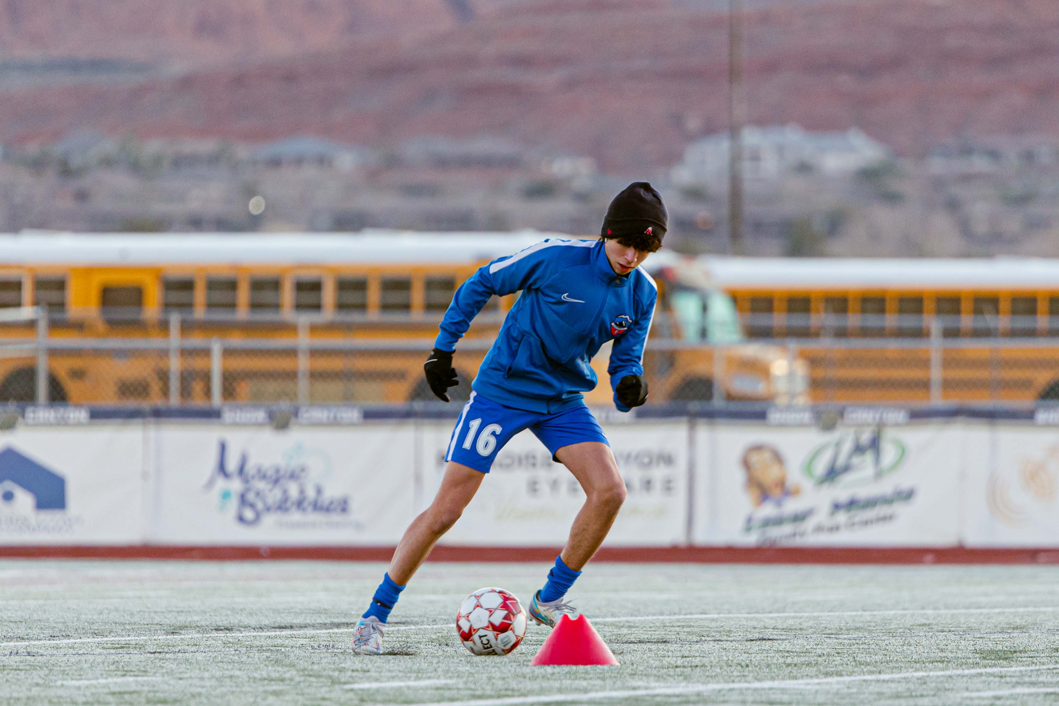 Player Dribbling Around A Cone Soccer player in a blue kit and beanie dribbles around a cone with school buses in the background
