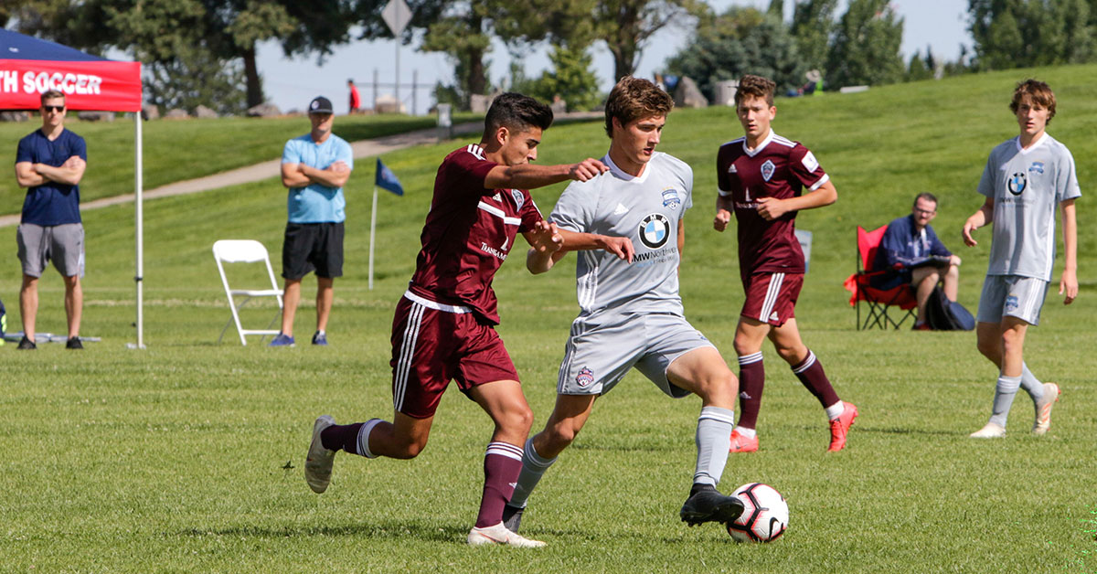 Boys Competing For The Ball Two youth players in gray and maroon kits compete for the ball as spectators watch