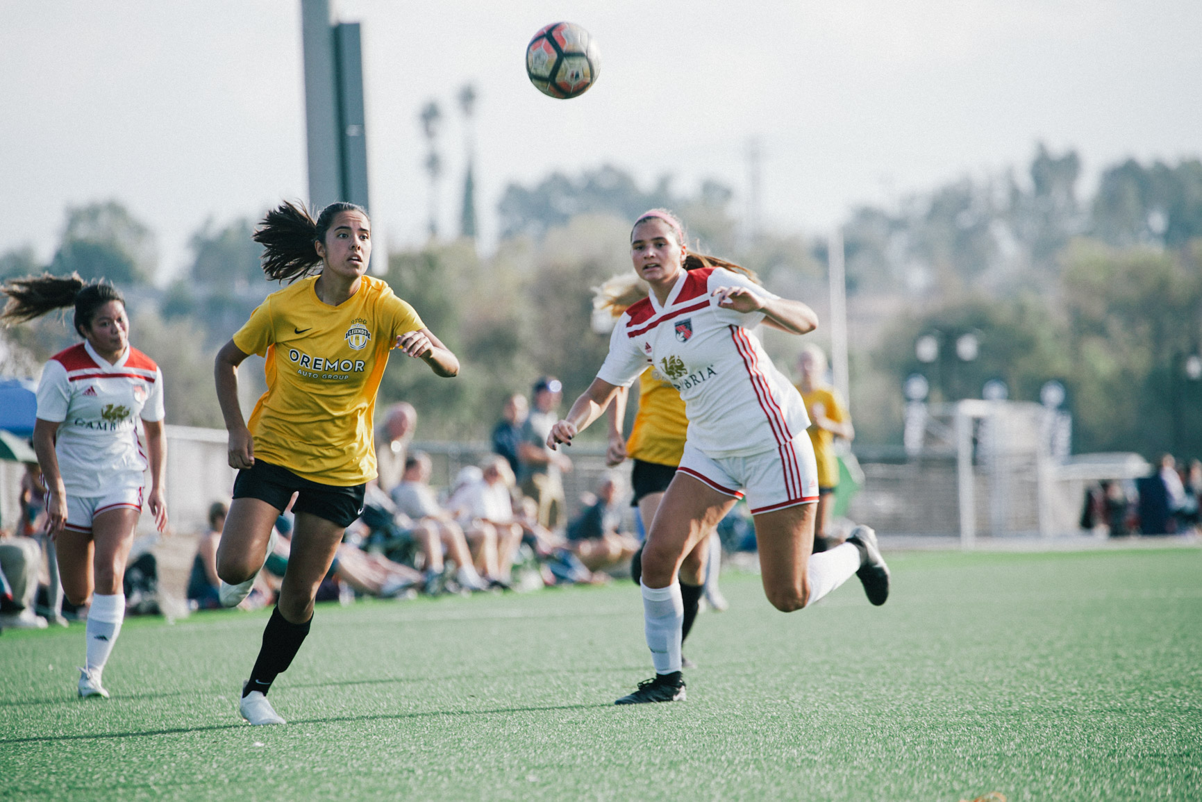 Two women players in yellow and white kits chase a ball in the air as spectators watch