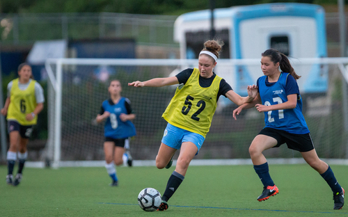 Female players in yellow and blue scrimmage bibs battling for the ball on a turf field