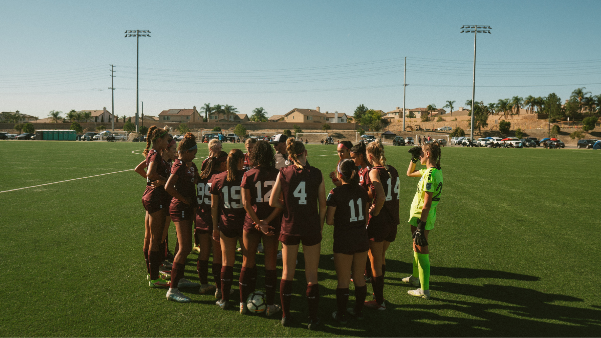 Girls Team Midfield Huddle Girls soccer team in maroon kits huddling together at midfield on a sunny turf field