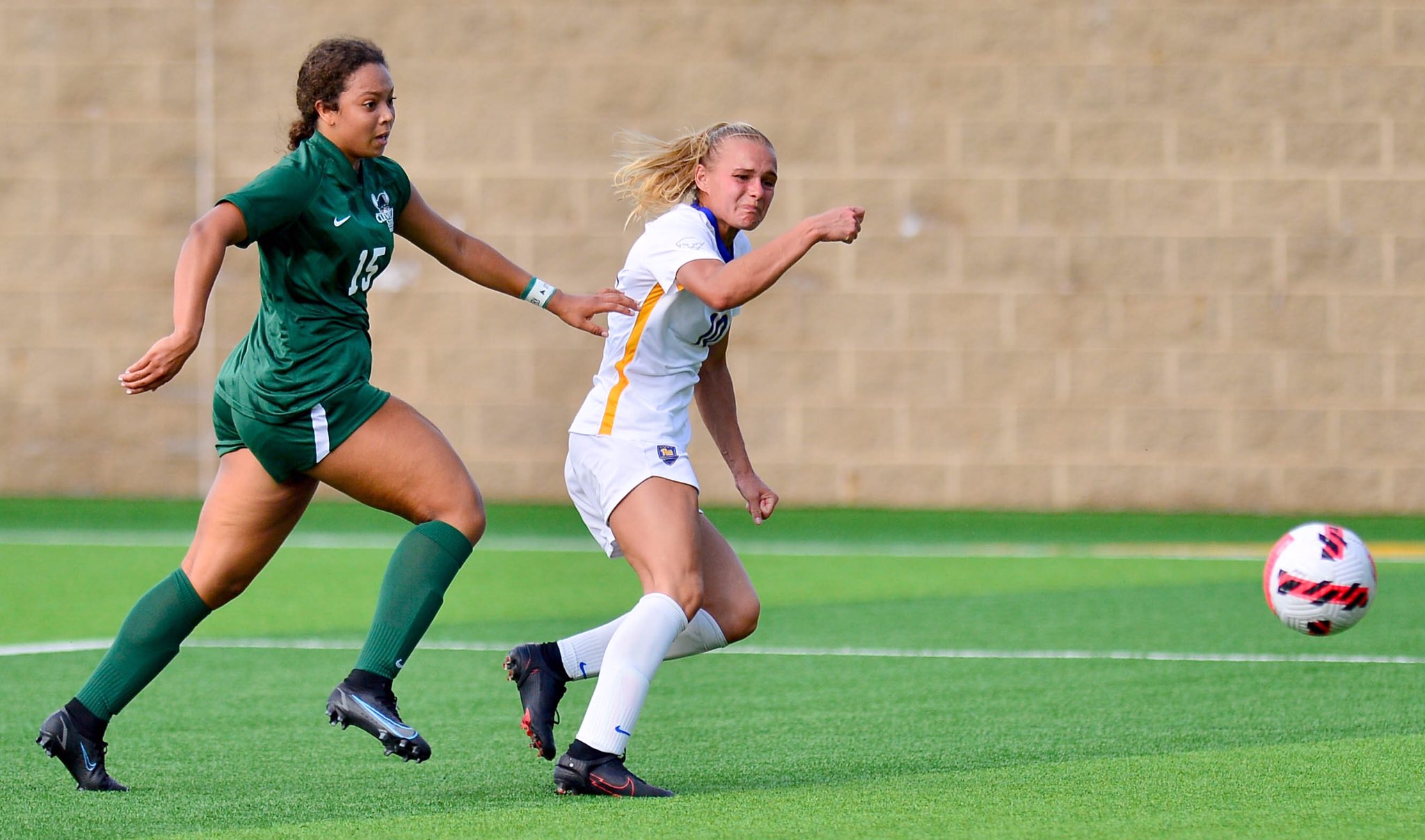 Female player in white kit shooting the ball past a defender in green during a college match
