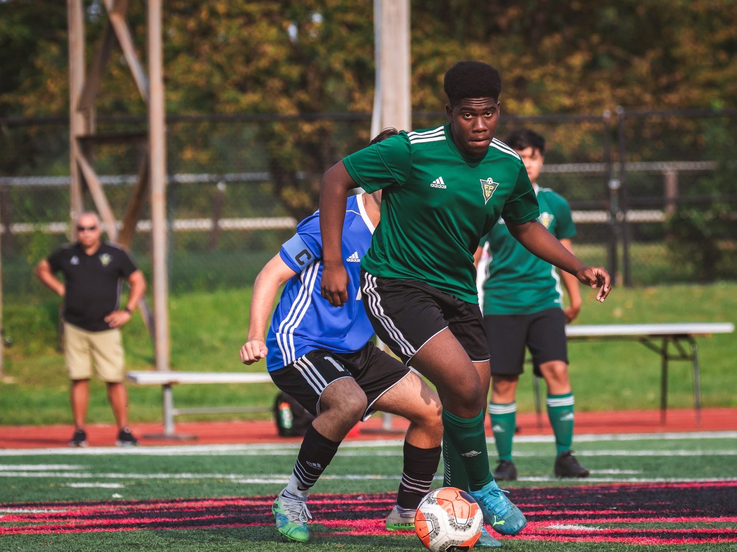 Player Shielding The Ball Youth player in green kit shielding the ball from a blue-shirted opponent on a turf field