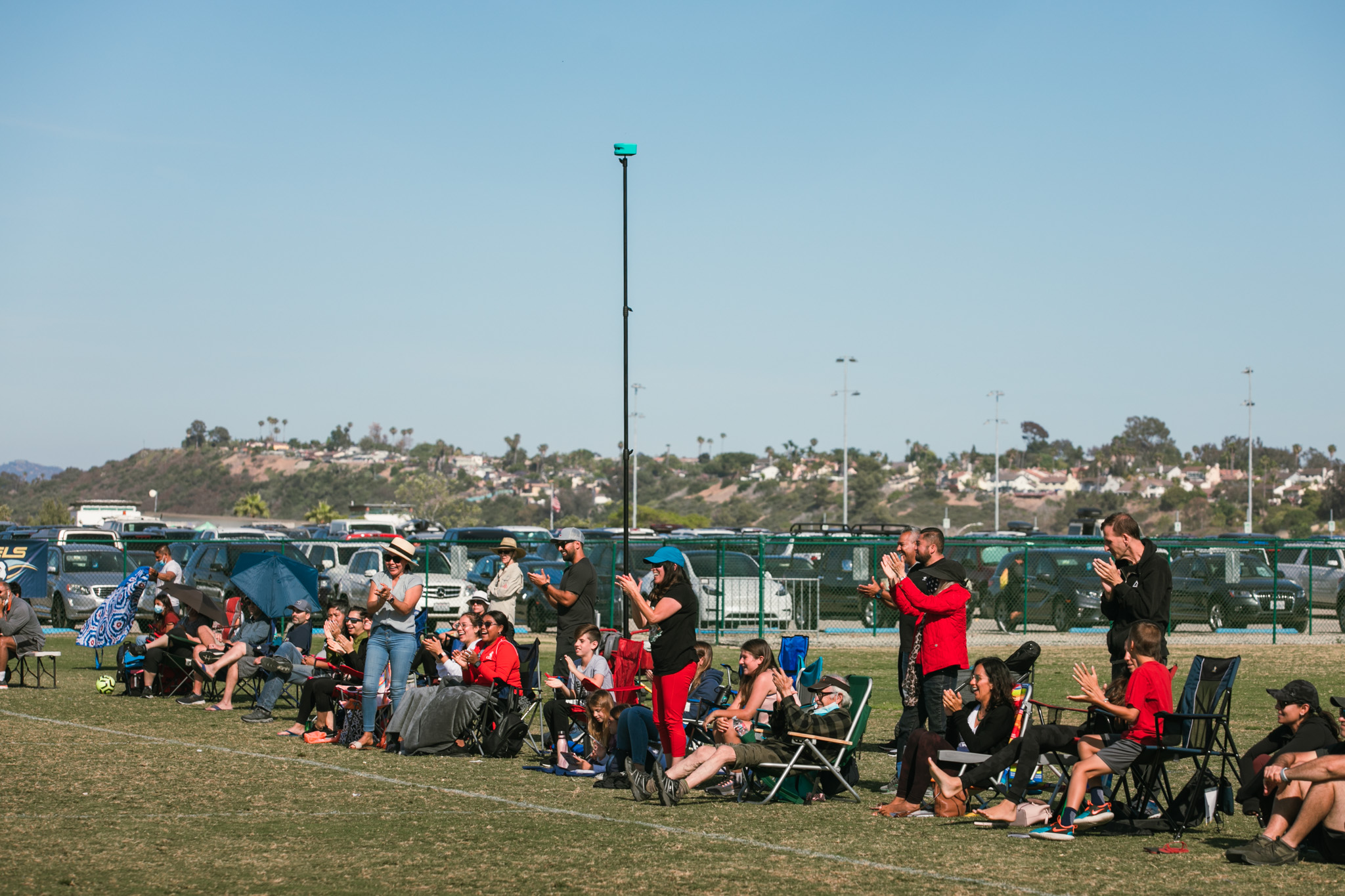 Spectators in camp chairs line a soccer sideline beneath a tall pole-mounted Trace camera.