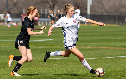 Female player in white FC Dallas kit winding up to strike past a defender in black