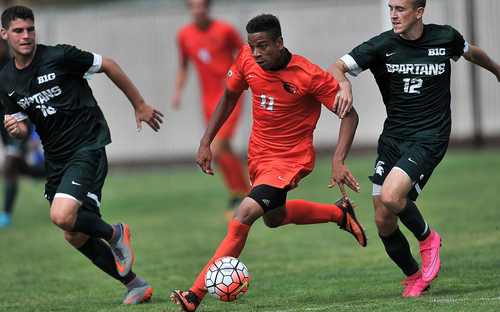 College Soccer Match Action College player in orange dribbles the ball between two Michigan State Spartans defenders in green.