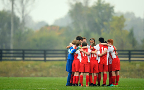 Youth soccer team in red jerseys huddles arm-in-arm together on a misty field.
