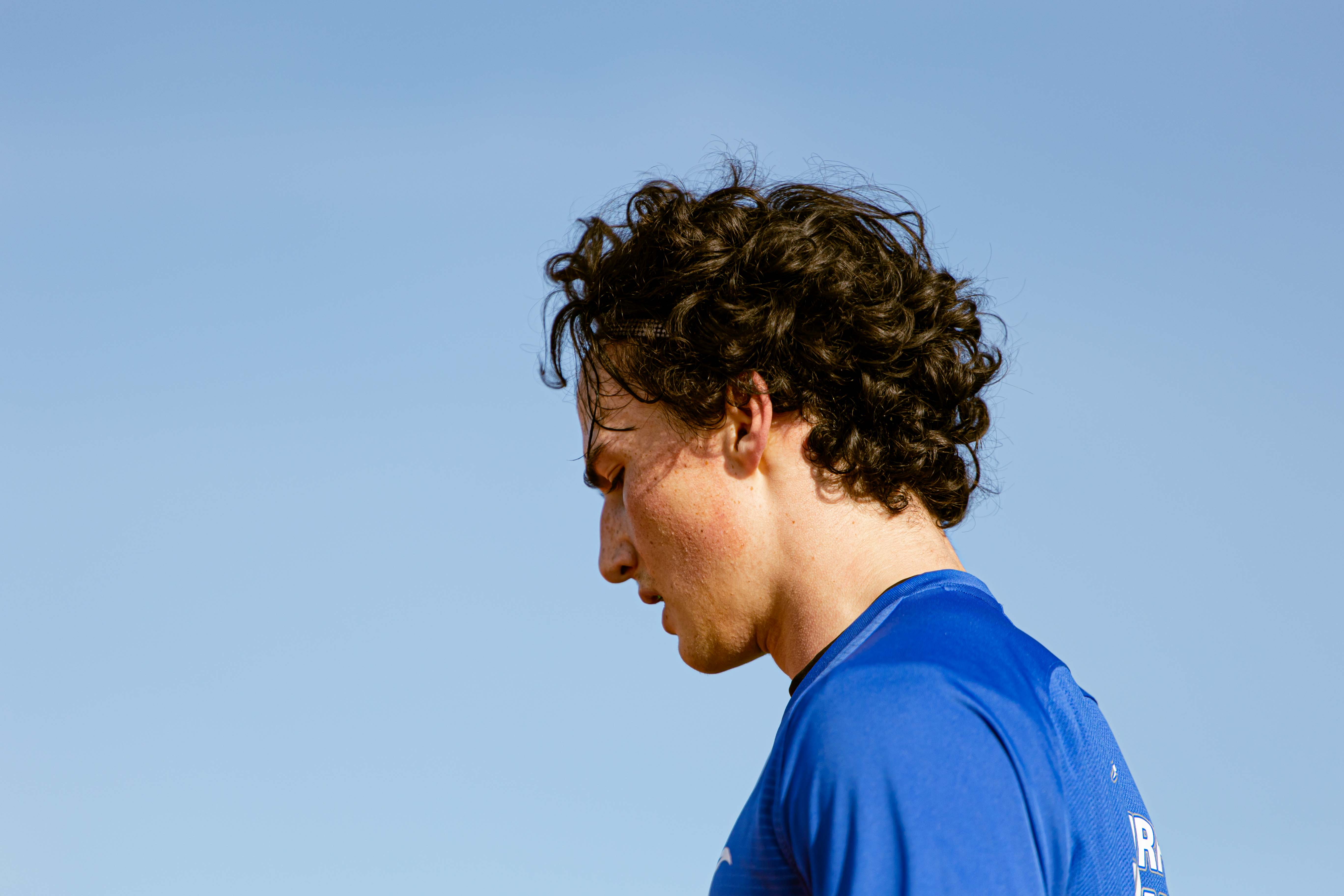 Close-up profile of a young soccer player in a blue jersey looking down against a blue sky.