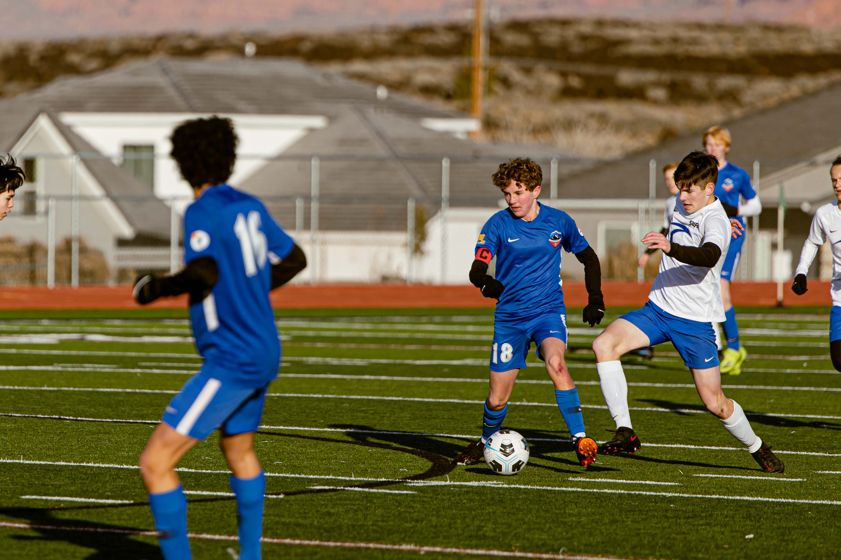 Youth Match Ball Battle Youth player in blue shields the ball from a white-jersey defender during an evening match.