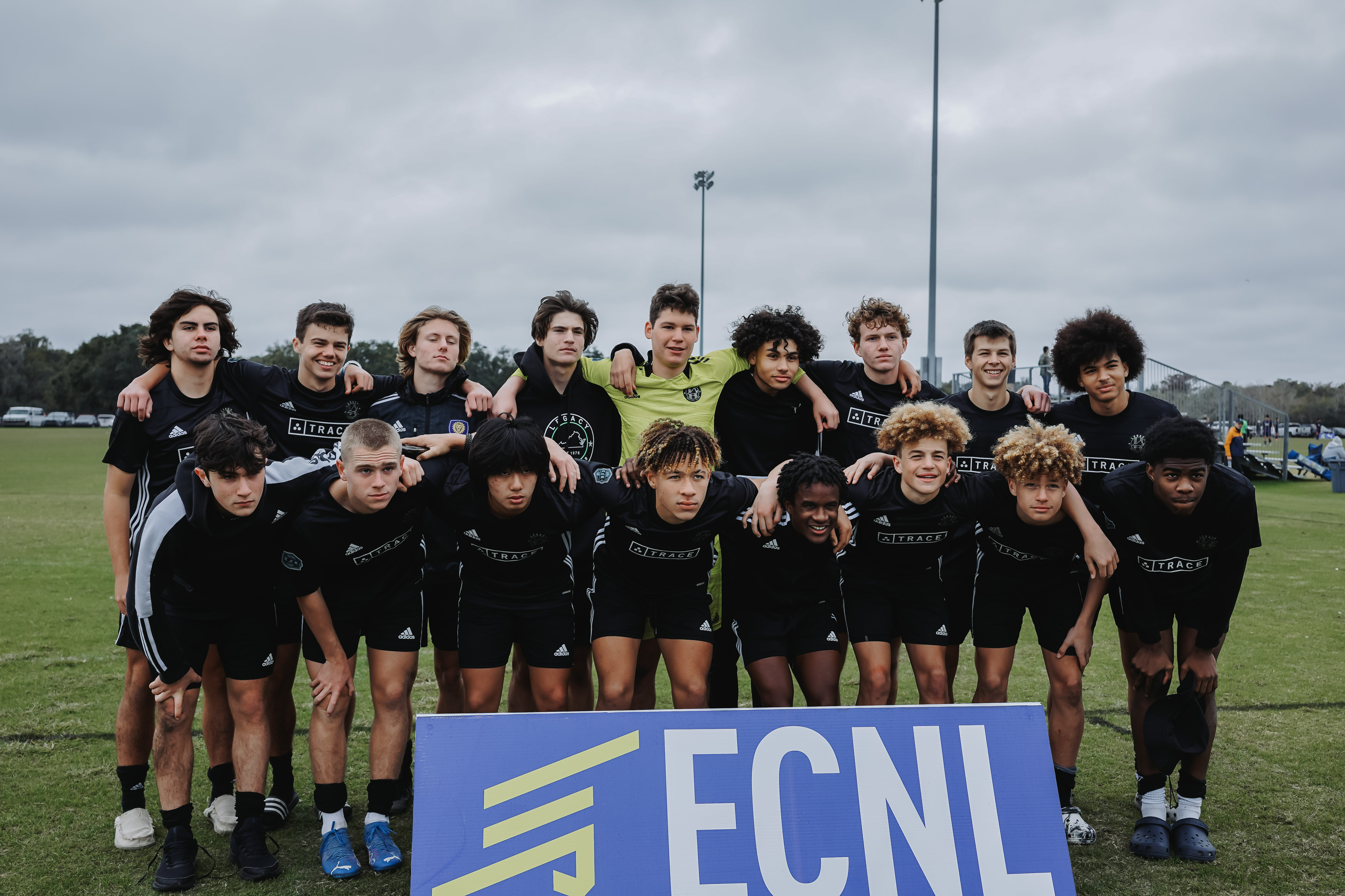 Boys soccer team in black uniforms posing together on a field behind a blue ECNL banner.
