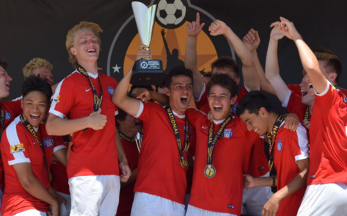 Youth players in red jerseys celebrate, holding a trophy overhead in front of a National banner.
