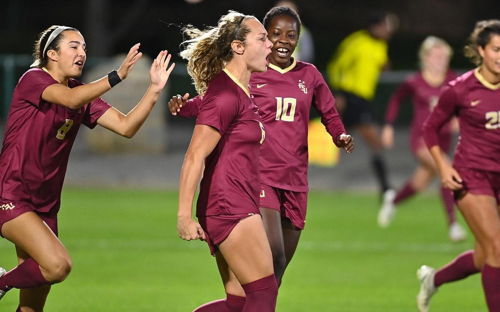 FSU Women Celebrating Goal Florida State women's players in maroon kits celebrate a goal on the field at night.
