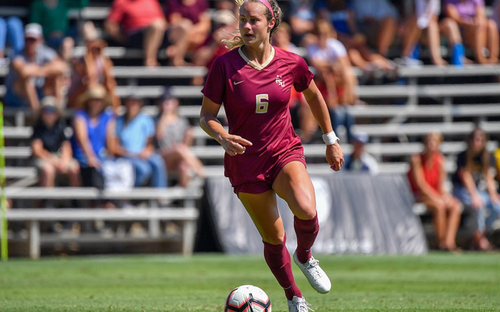 Female college soccer player in a maroon number 6 kit dribbles the ball past stadium bleachers.