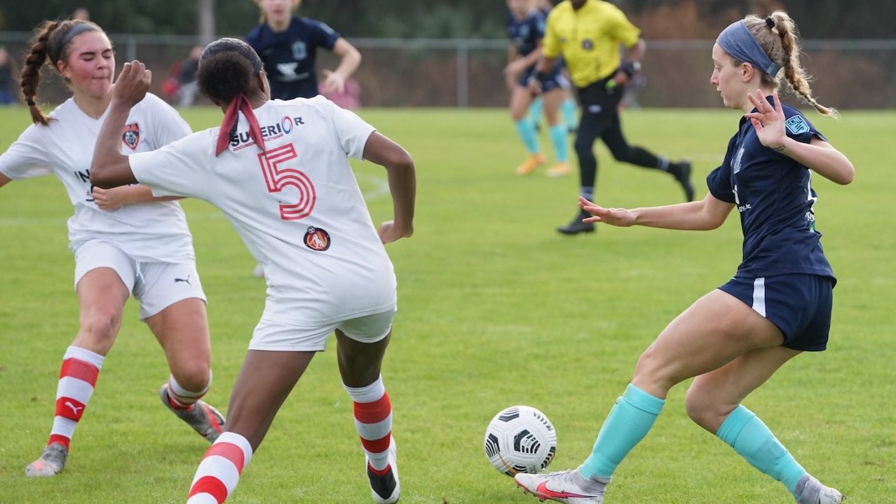 Youth girls compete for the ball, a navy-clad player facing two white-and-red opponents during a match.