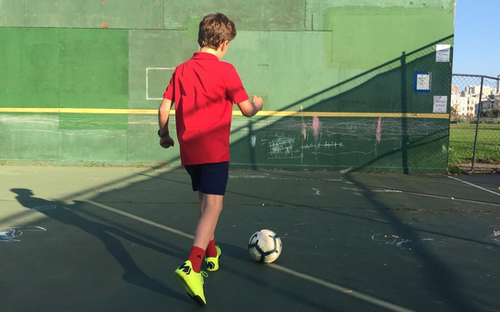 Boy in a red shirt dribbling a soccer ball against a green handball wall on an outdoor court.