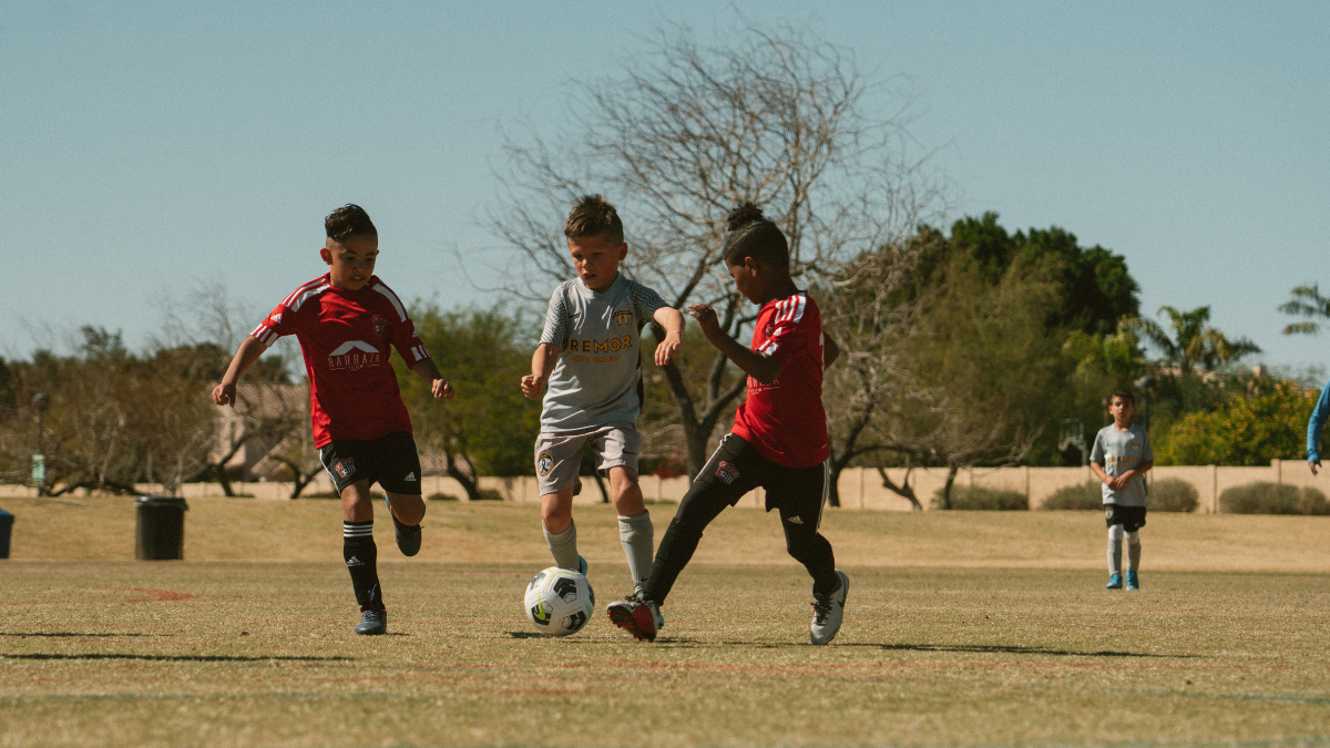 Young Boys Battle For The Ball Three young boys in red and grey kits battle for the ball on a dry grass field lined with trees.