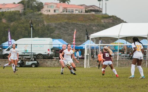 Girls soccer match at an outdoor showcase with tournament tents and a hillside home behind the field.