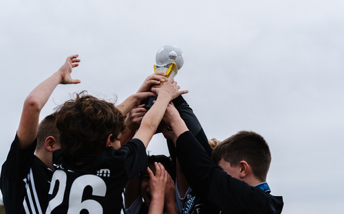 Youth boys in black kits raise a trophy topped with a soccer ball against a grey sky.