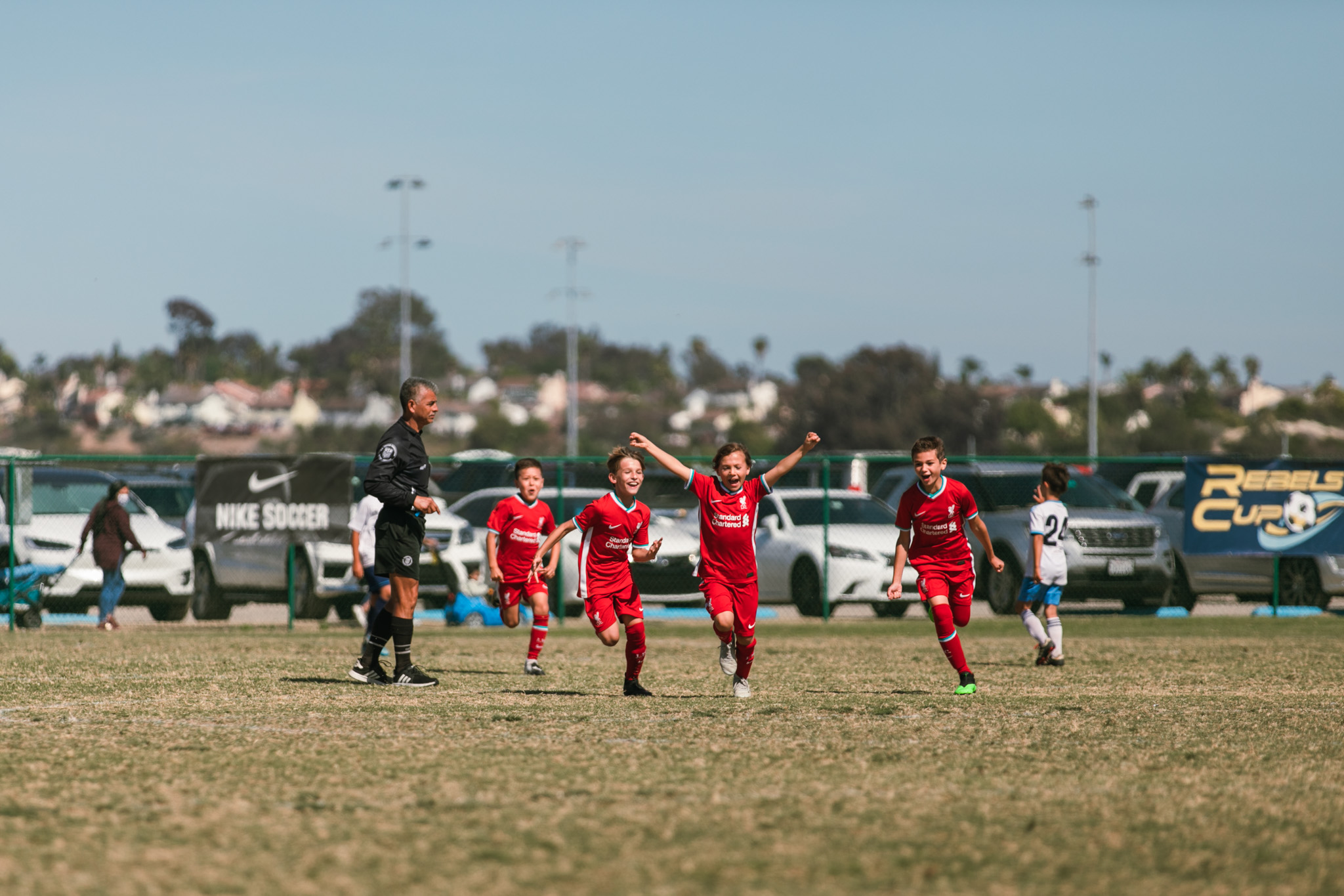Young boys in red kits run and celebrate with raised arms as a referee looks on at a tournament.