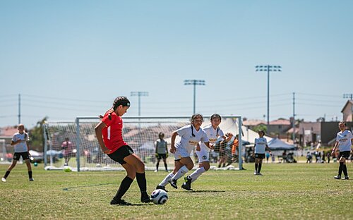 Girl Attacking The Defense Girl in a red number 1 kit dribbles toward two white-kitted defenders on a sunny tournament field.
