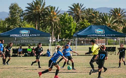Girls Match At Silverlakes Girls in blue and black kits compete for the ball at Silverlakes with palm trees and event tents behind.