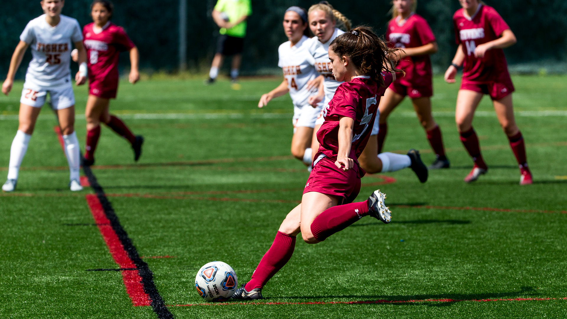 College woman in maroon winds up to strike the ball as opponents in white and maroon converge.