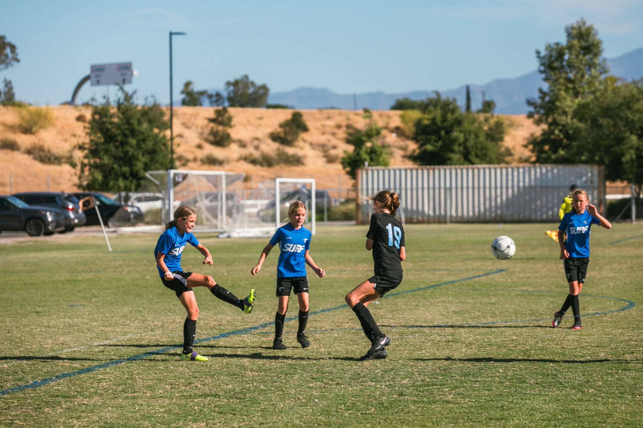 Girls Surf Match On Grass Girls in blue Surf kits and black kits contest the ball on a grass field backed by dry hills.