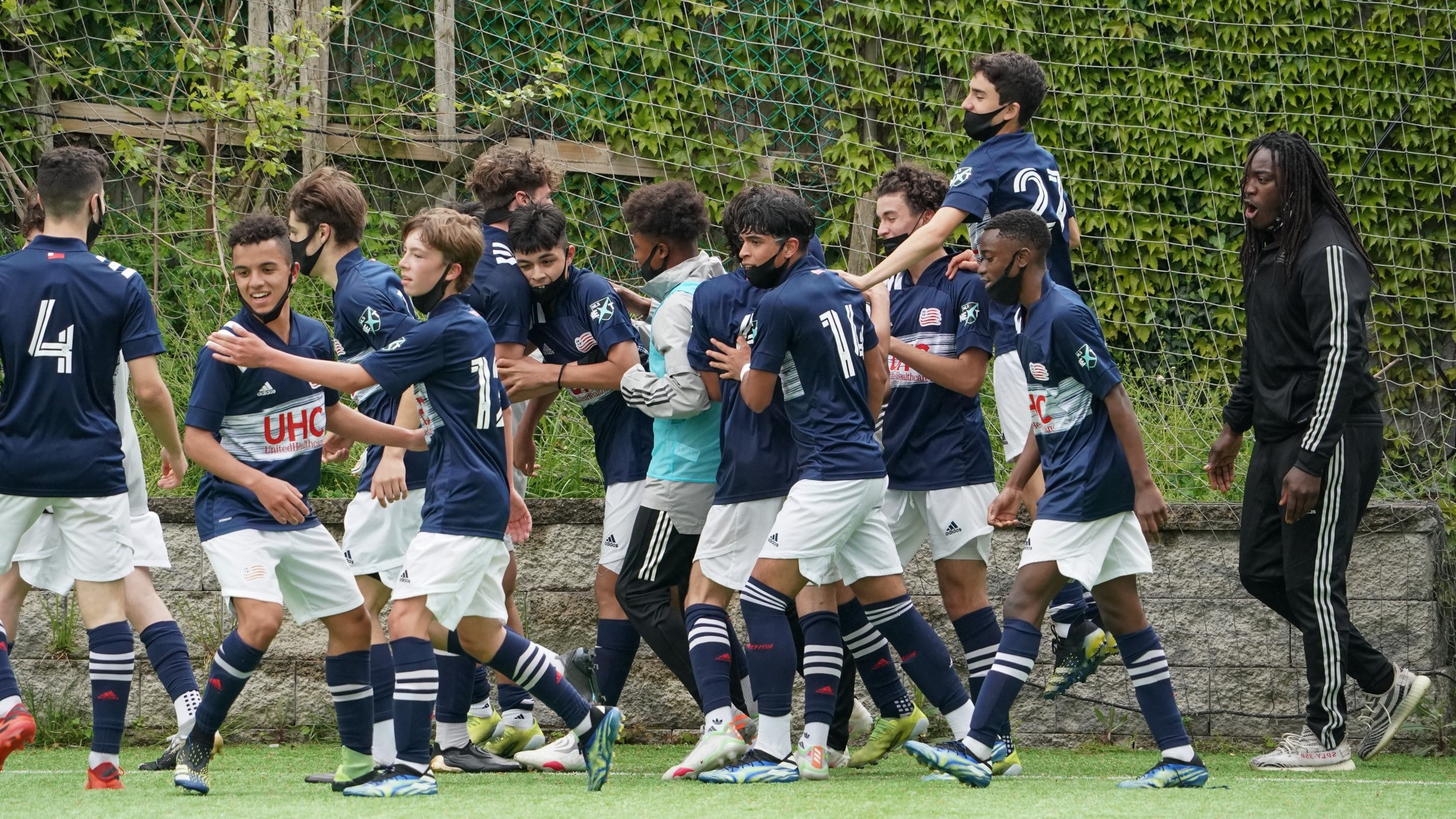 Team Celebrates A Goal Teen boys in navy kits swarm together celebrating a goal beside the net as a coach cheers.