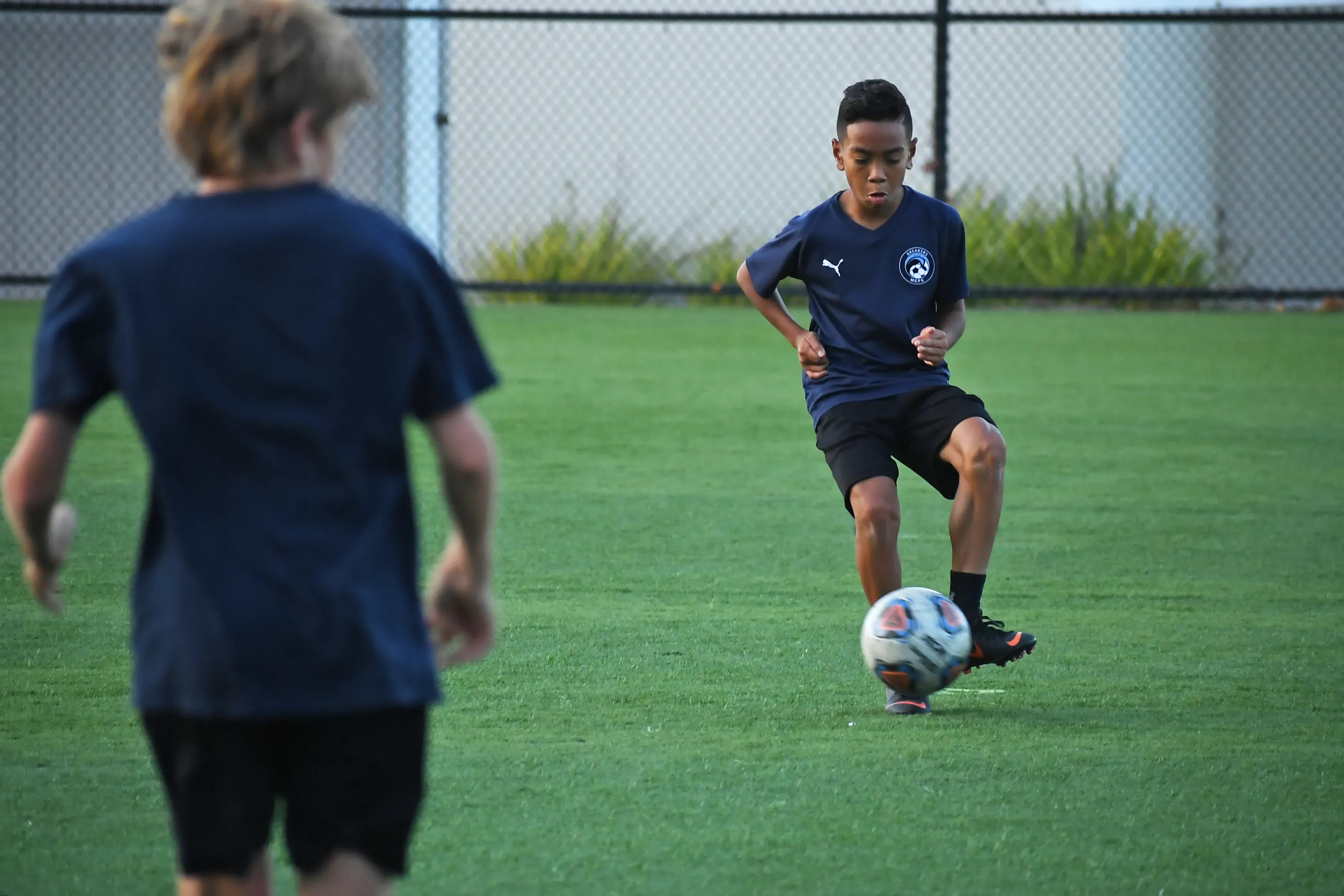 Teen boy in navy controls a soccer ball with his knee on a turf field as a teammate watches.