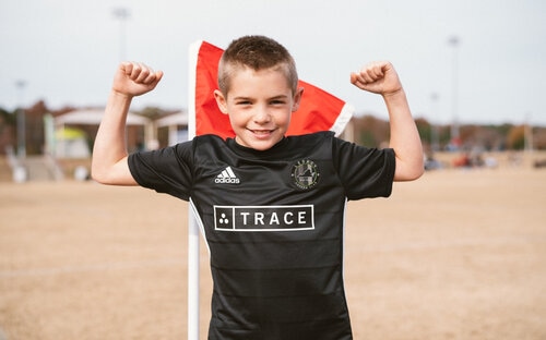 Young Player Flexing Arms Smiling young boy in a black Trace soccer jersey flexing both arms beside a red corner flag