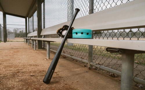 Trace Camera In Dugout Baseball bat and teal Trace camera unit resting on a metal dugout bench beside a field