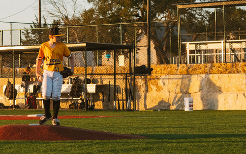 Youth baseball pitcher in a gold jersey standing on the mound at golden hour