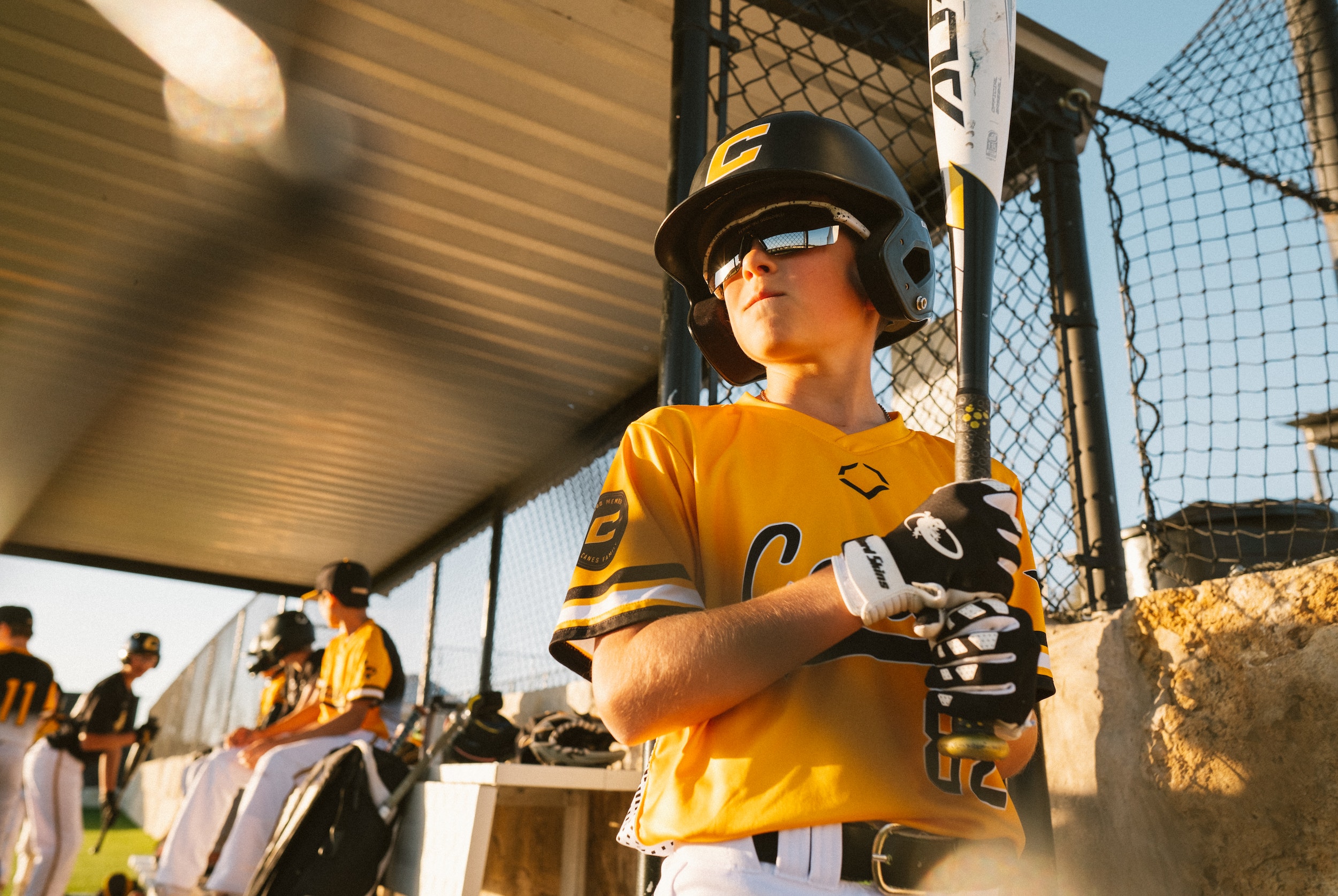 Batter Waiting In Dugout Young batter in a gold jersey and helmet holding a bat in the dugout at golden hour