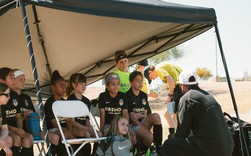 Youth soccer players seated under a shade tent listening to a coach before a game
