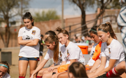 Girls in white soccer jerseys resting on a sideline bench, one standing with arms crossed looking down