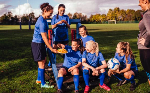 Youth girls soccer team in blue kits sharing a plate of orange slices on the field with their coach
