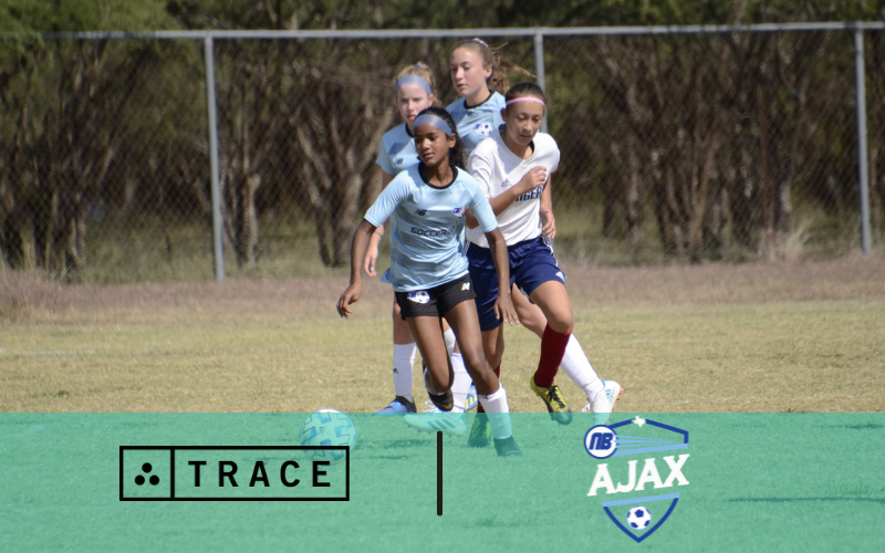 Youth girls soccer players competing for the ball on a field; Trace and NB Ajax logos