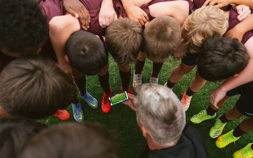 Overhead view of a youth soccer team huddled around a coach showing them a phone screen